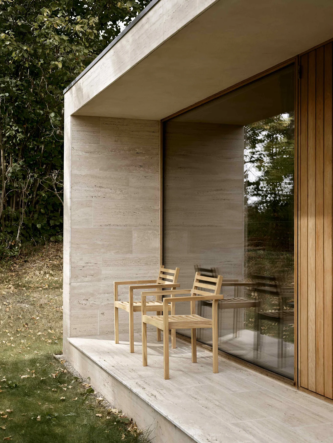 To lyse trelamellstoler på en beige steinterrasse ved en moderne bygning med glassvindu og trepanel.