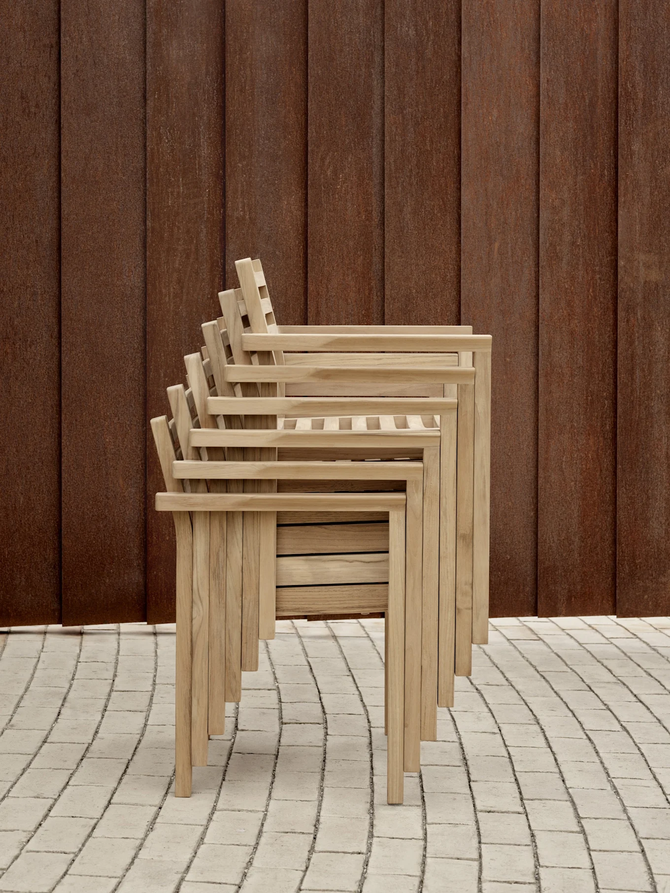 Stabel med fem lyse trehagerstoler med lamellrygg og sete, foran en rustbrun vegg på et flislagt gulv.