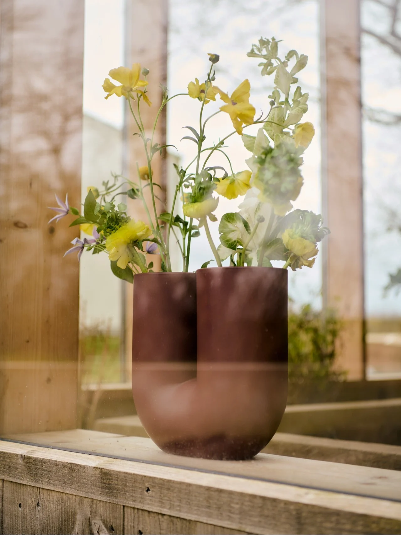 Mørkebrun U-formet vase med gule, lysegrønne og lilla blomster på en trevinduskarm.