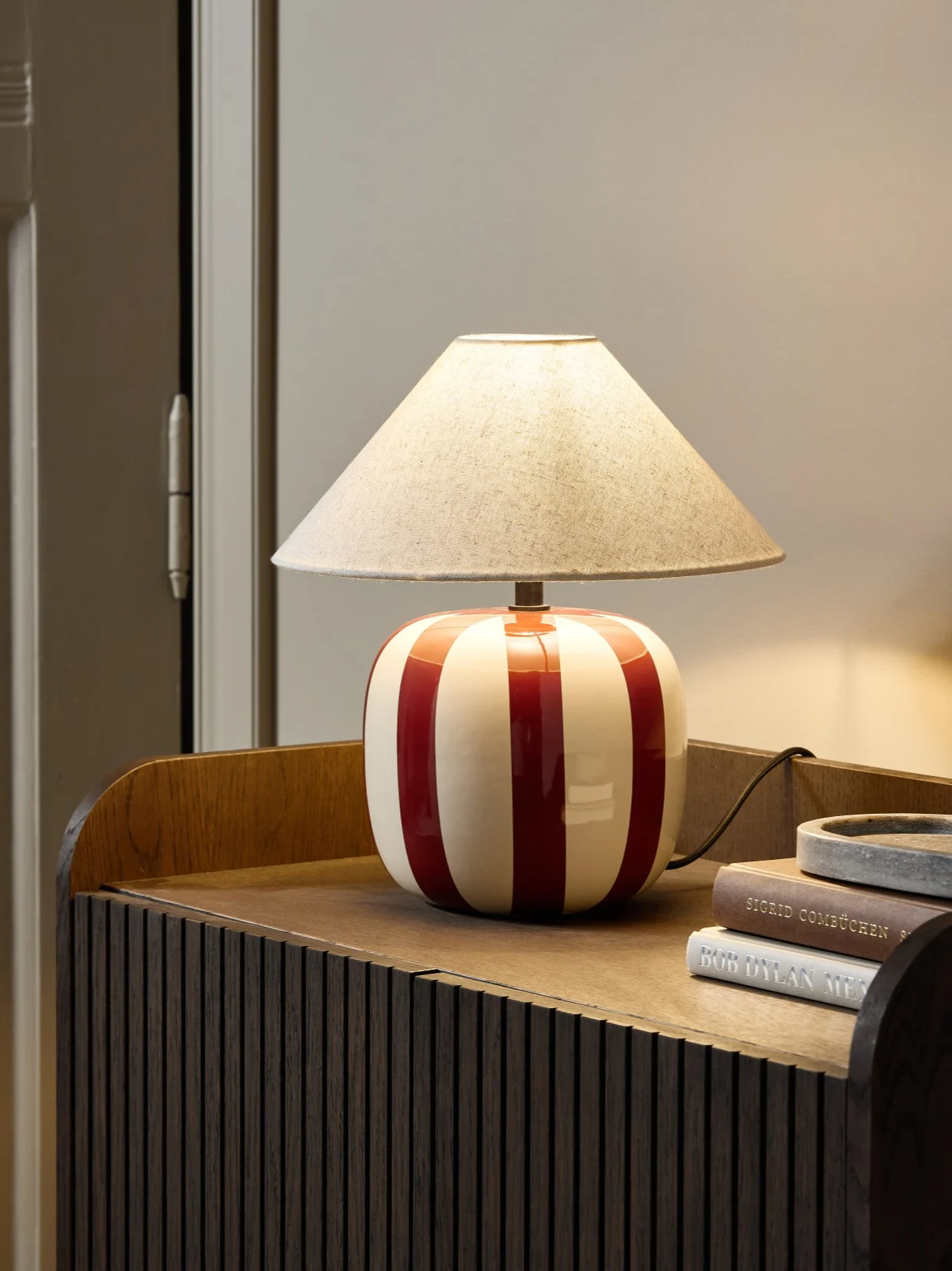 En bordlampe med rød- og hvitstripet keramikkbase og beige tekstilskjerm står på et mørkt treskap.