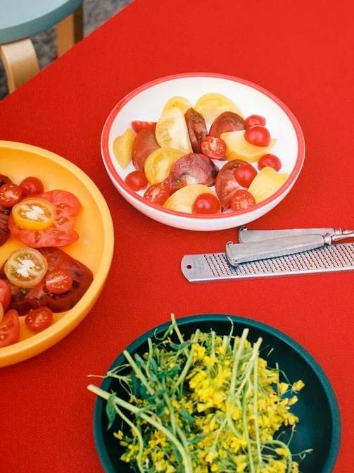 Fersk tomatsalat med gule og røde tomater, en ostehøvel og en bolle med grønne skudd og gule blomster på et rødt bord.