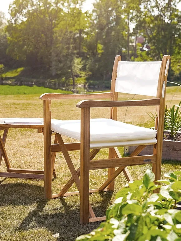 Regissørstol i teak med hvitt stoff og pute, ved siden av et matchende lite bord utendørs i en hage.