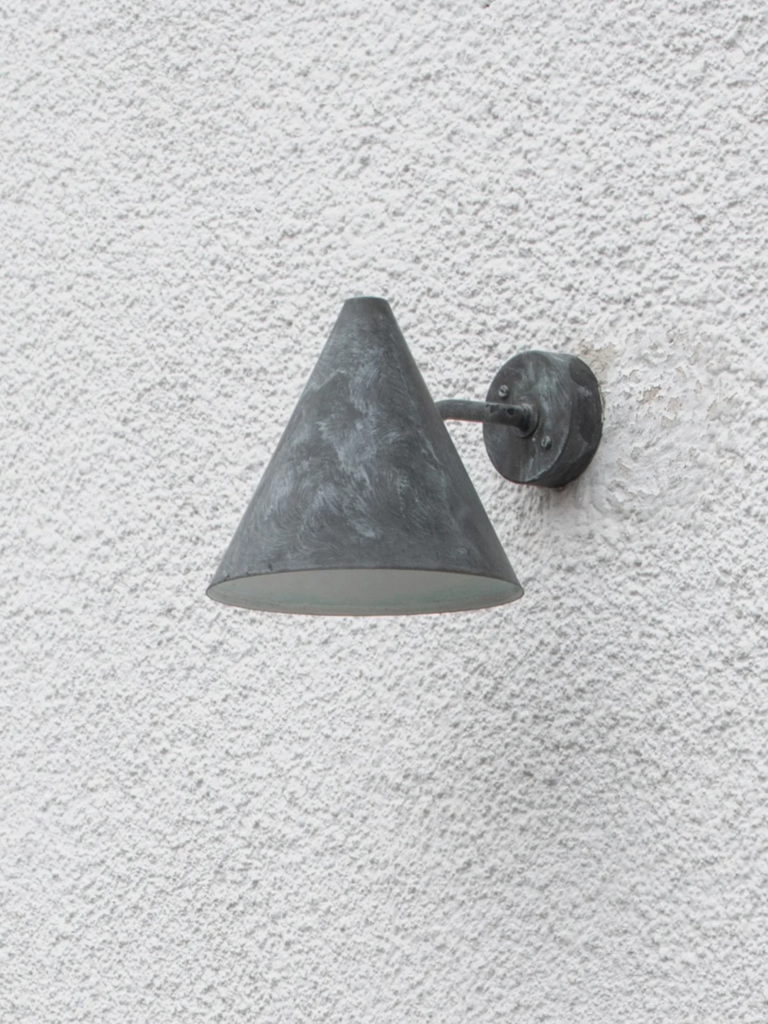 En forvitret mørkegrå konisk utendørs vegglampe montert på en teksturert hvit vegg.