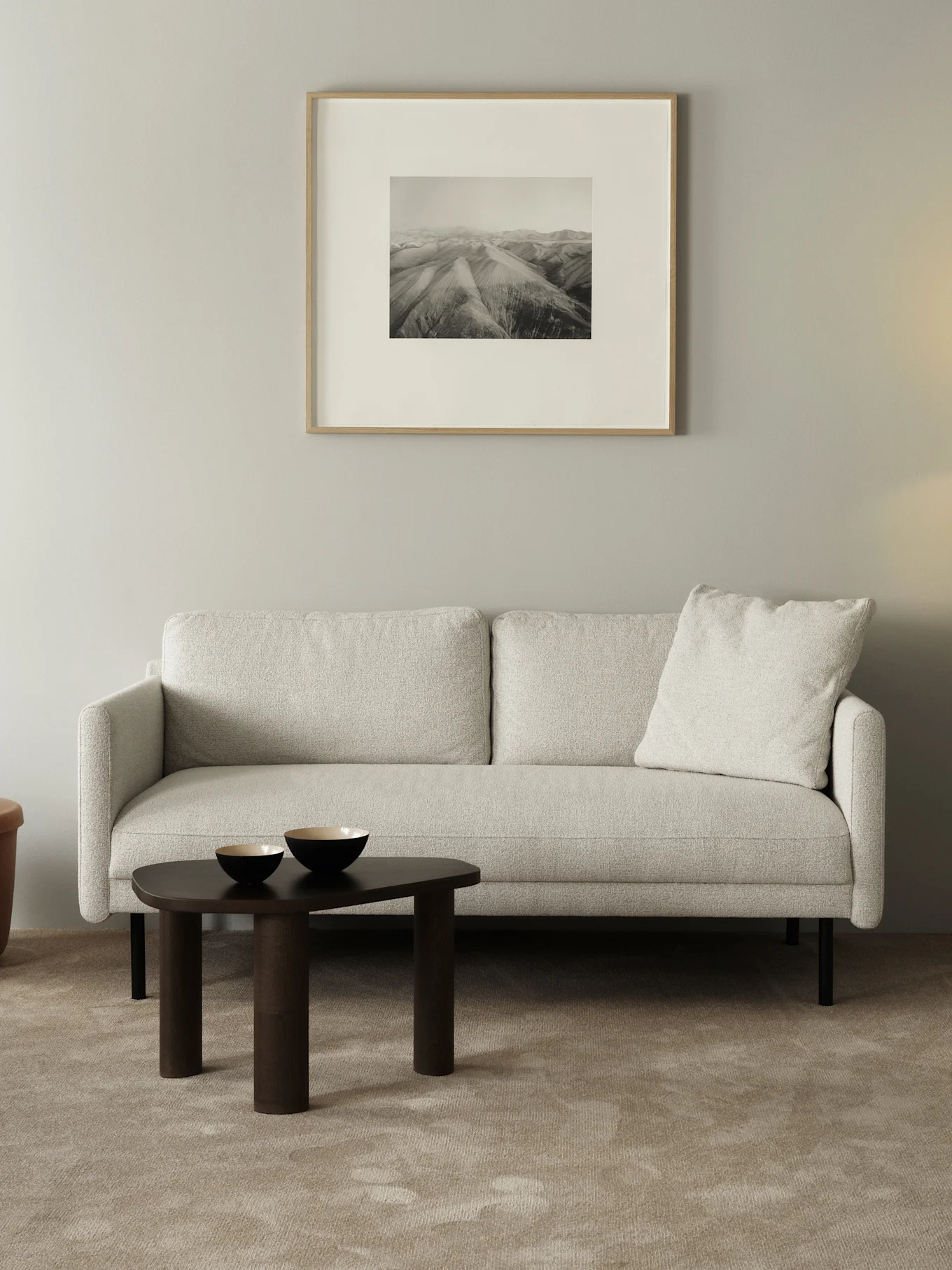 Lys teksturert sofa, mørkt organisk salongbord med to gullkantede skåler, innrammet svart-hvitt fjellkunst.