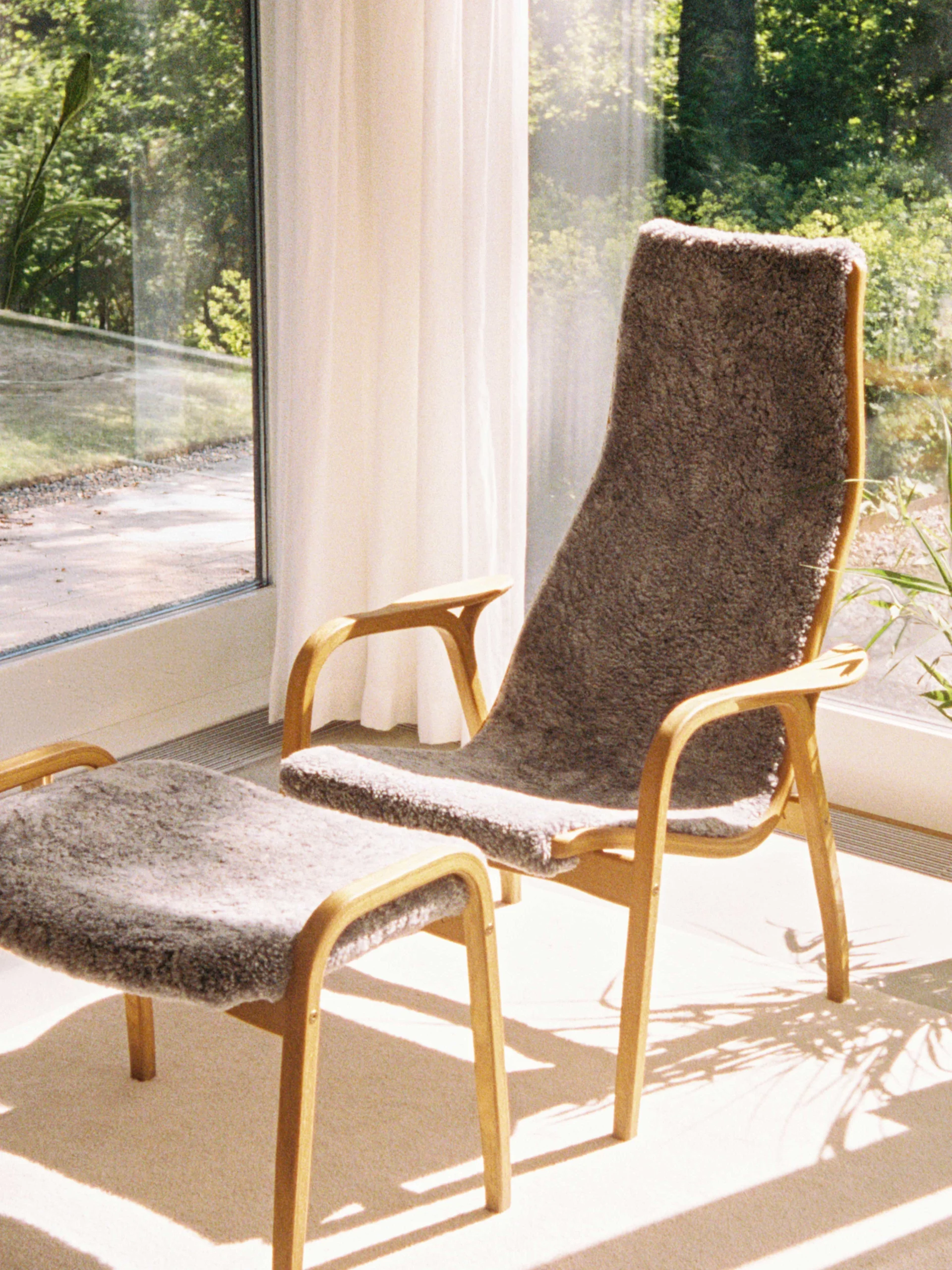 Høybent lenestol med brunt saueskinn og elefantpute, matchende fotskammel og sfærisk papirlampe.