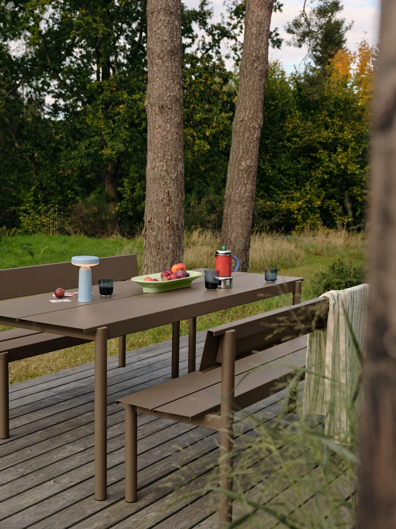 Utendørs piknikbord med lyseblå lampe, frukt og rød termos på en treplatting.