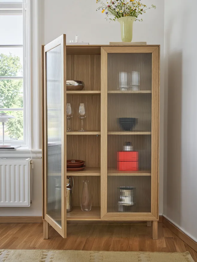 Lyst eikeskap med riflede glassdører, den ene åpen, viser servise og glass. Blomster i vase på toppen.