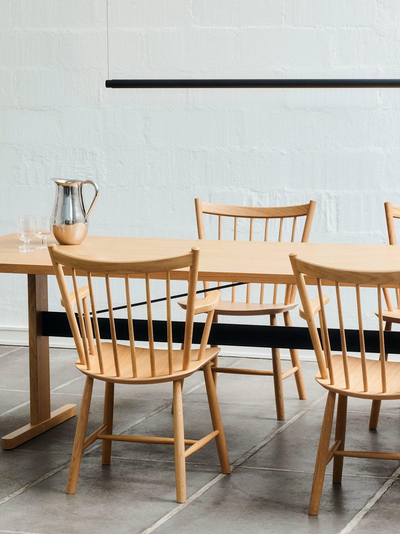 Et spisebord i lyst tre med matchende stoler med spiler, en sølvkanne og to glass. En svart lineær lampe henger over.