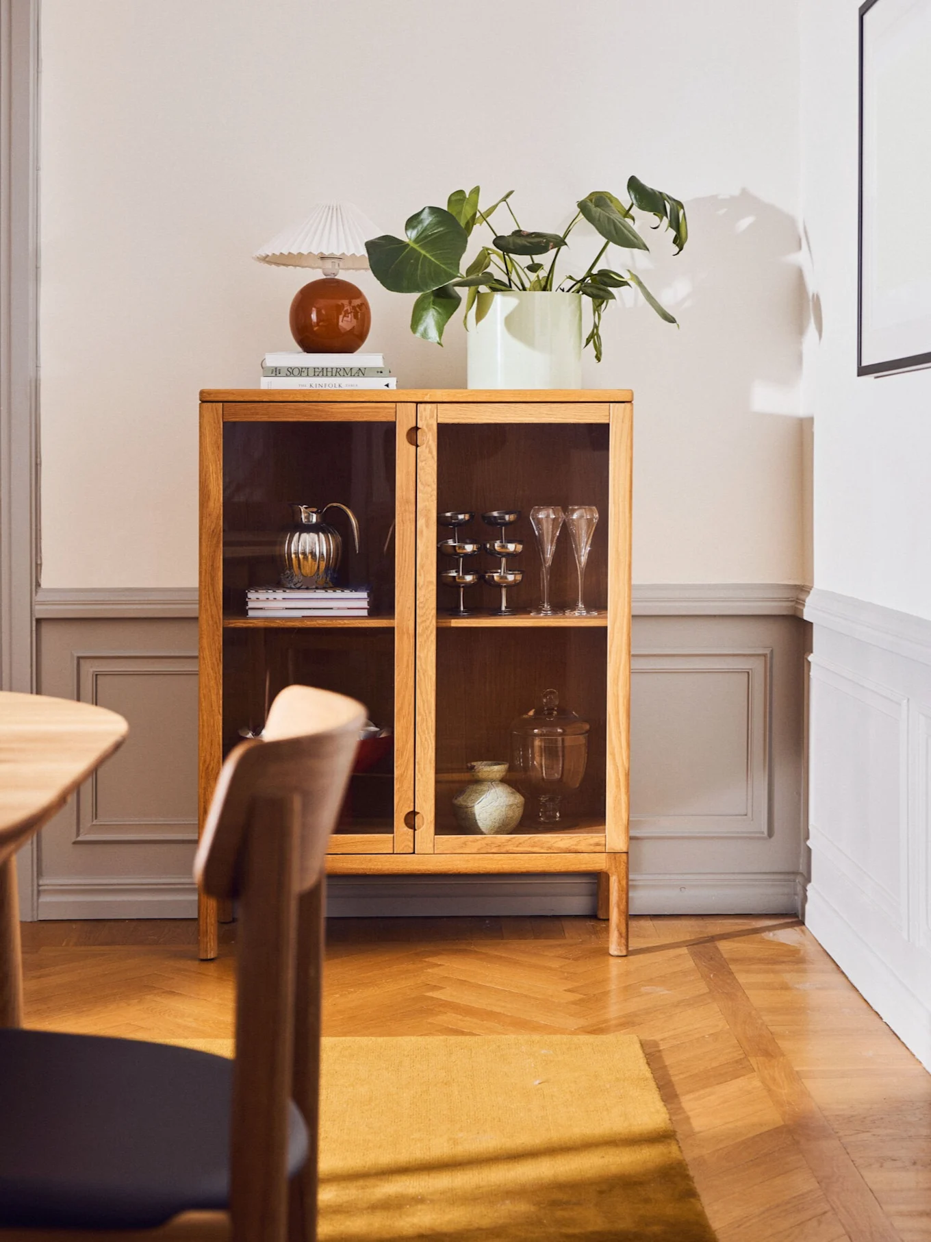 Eiketreskap med glassdører, interiørgjenstander, en lampe, bøker og en plante. Rommet har tregulv og et gult teppe.