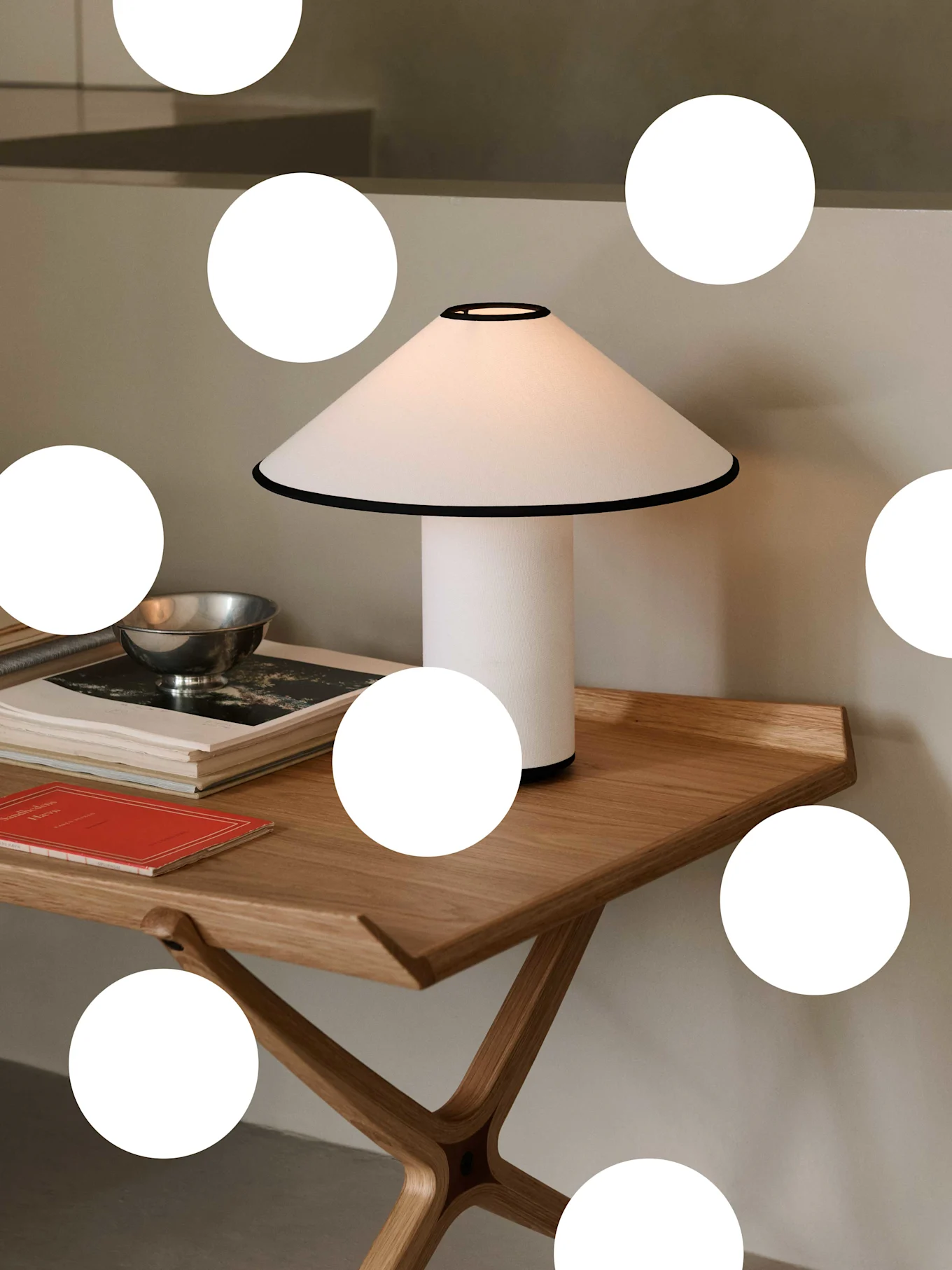 Hvit bordlampe med konisk skjerm og svart kant på et lyst trebord, med bøker og en sølvbolle.
