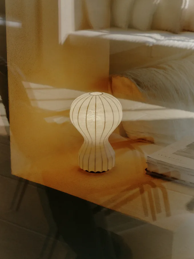 En lys, ribbet, timeglassformet bordlampe på et treunderlag med et soverom i bakgrunnen.