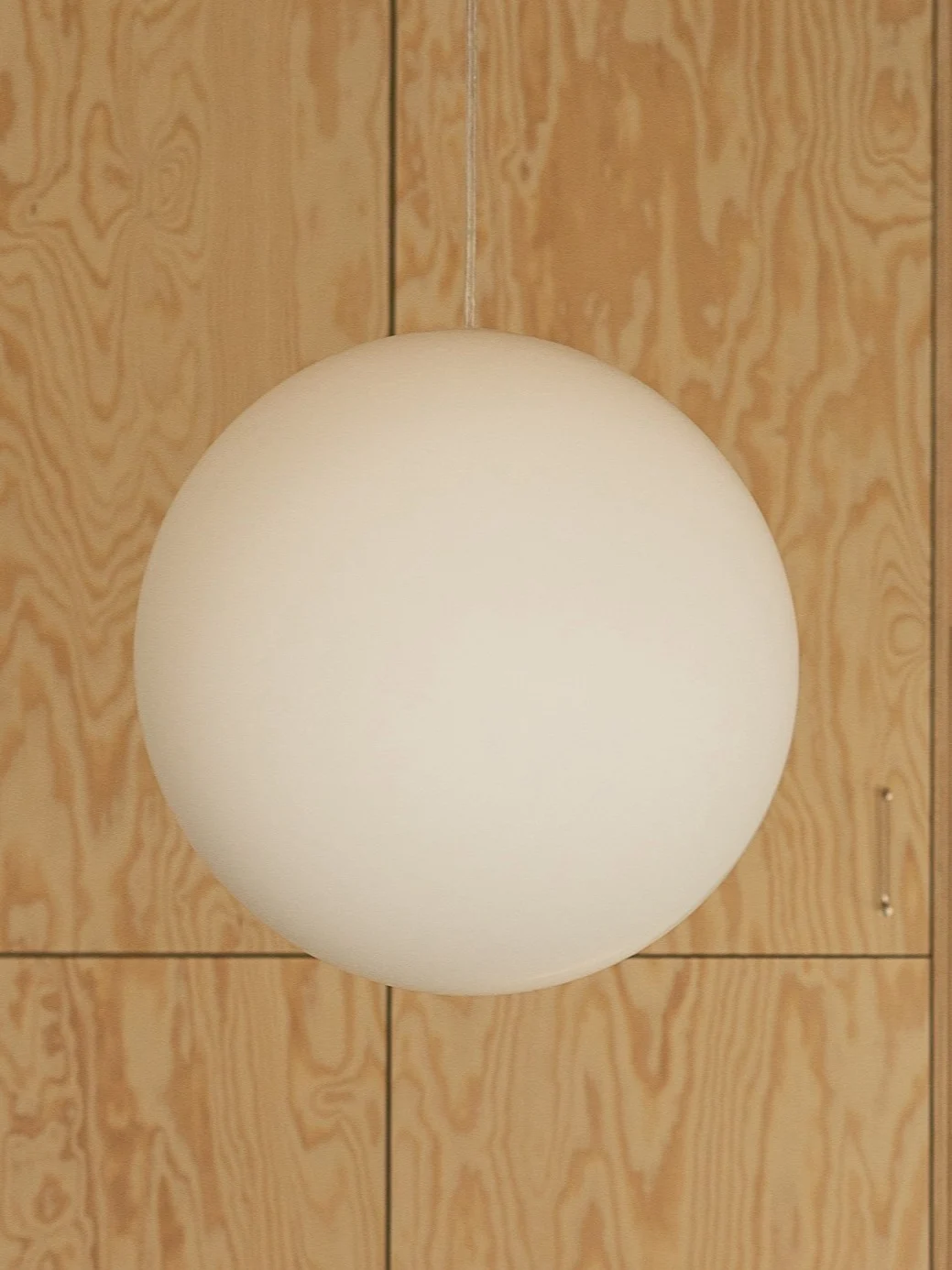 En hvit, sfærisk pendellampe henger foran en lys trevegg med synlig årring.
