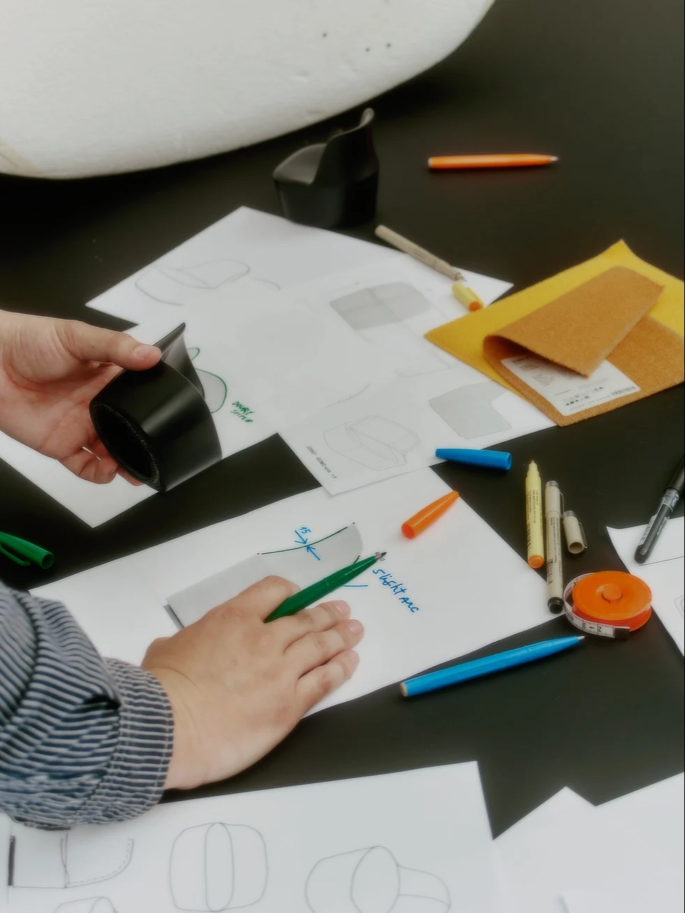 Hender skisserer og holder en svart prototype blant designpapirer, stoffprøver og penner på et mørkt bord.