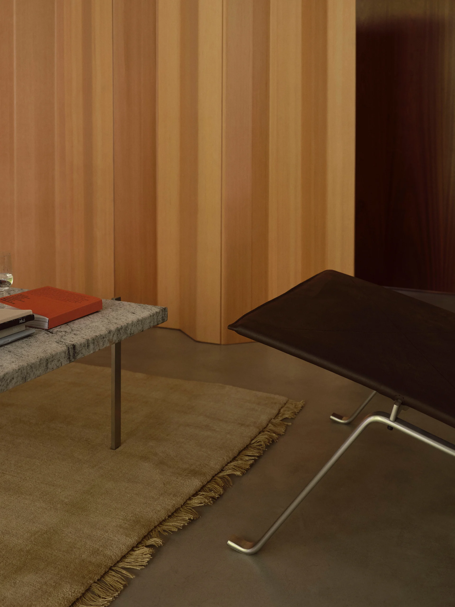 Interiør med et salongbord i granitt med en oransje bok, et beige teppe og en mørkebrun skinnlenestol mot en lys trepanelvegg.