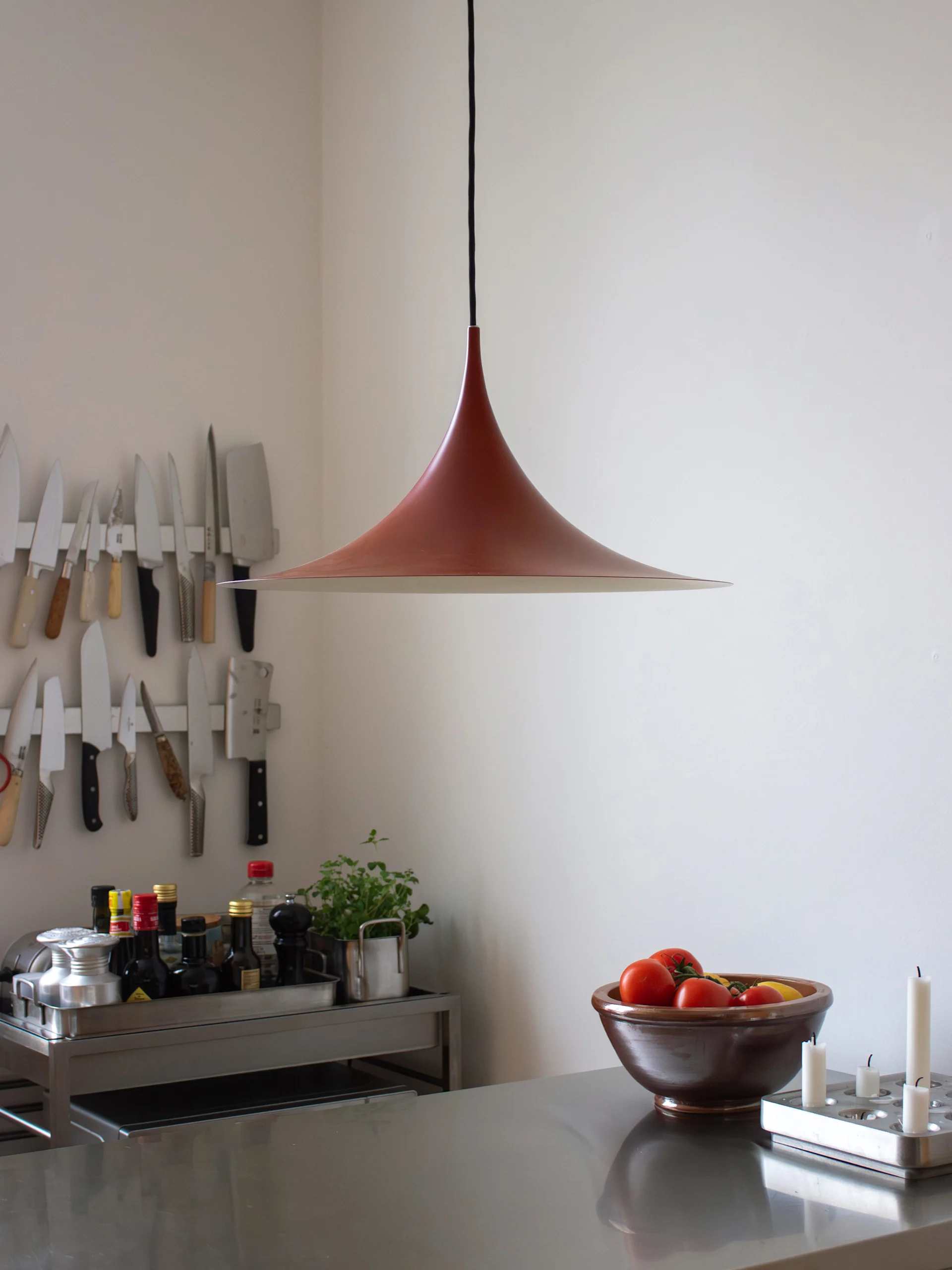 Moderne kjøkken: rødbrun klokkeformet lampe, benkeplate i rustfritt stål med tomater og lys, vegg med hengende kniver.