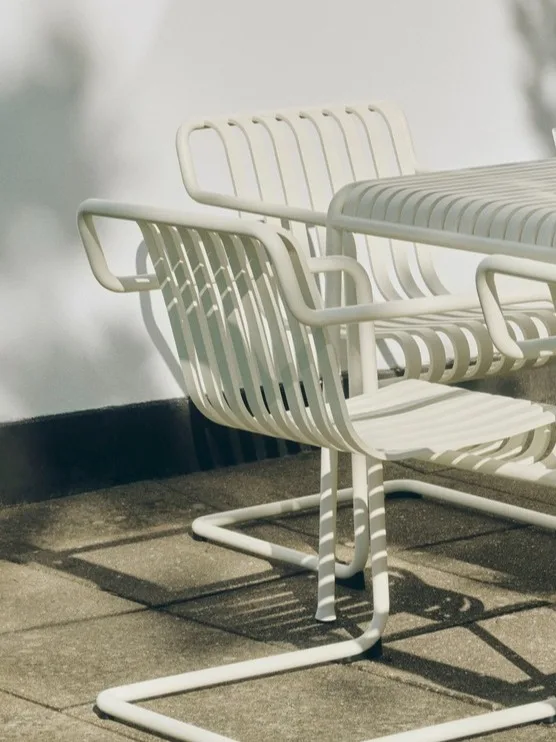 To hvite utestoler i metall med lameller og et matchende bord på en uteplass i sollys.