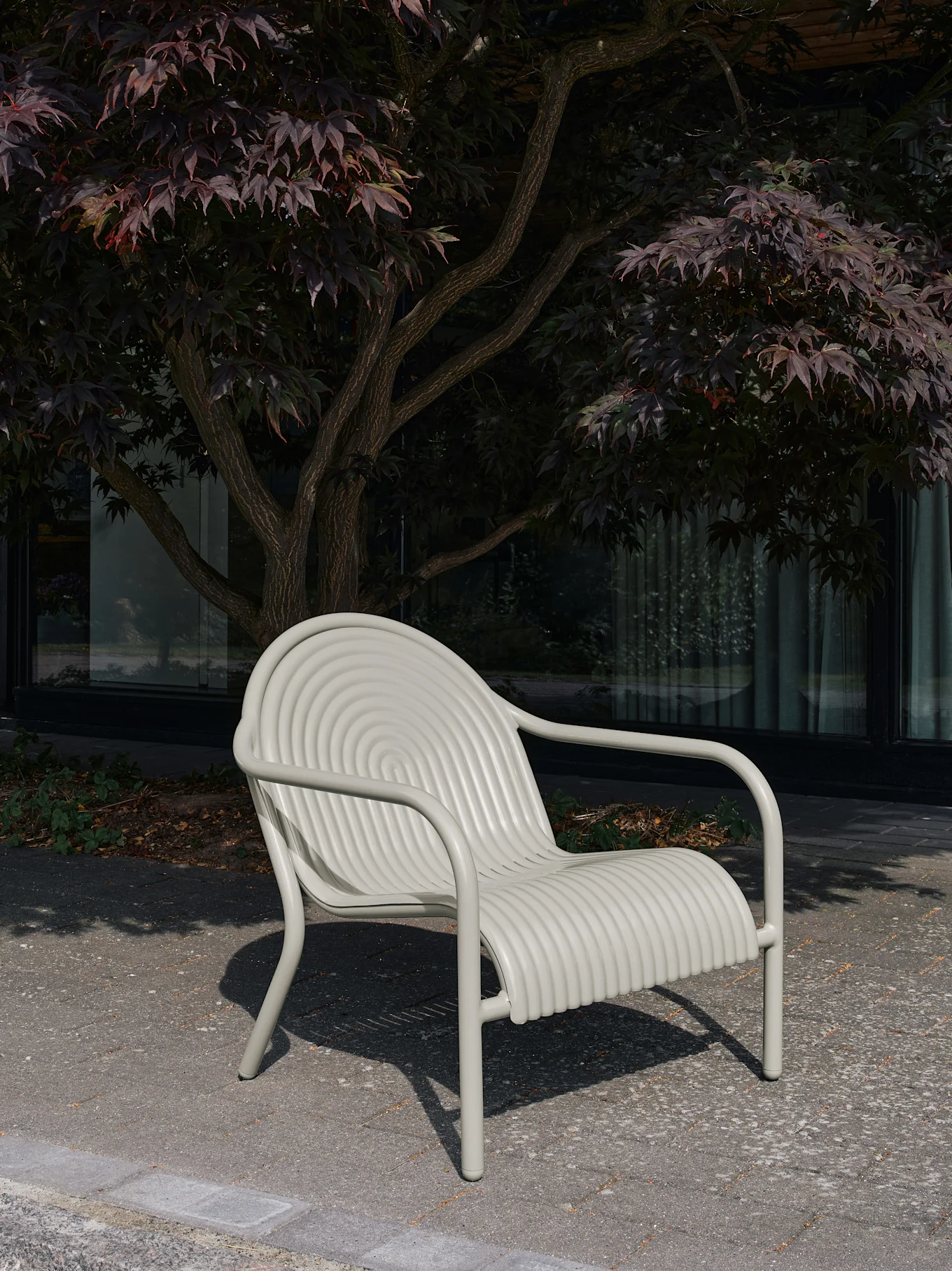 En lys grå utendørsstol med ryggstøtte med konsentriske sirkler og sete med horisontale riller, plassert på heller med et tre bak.