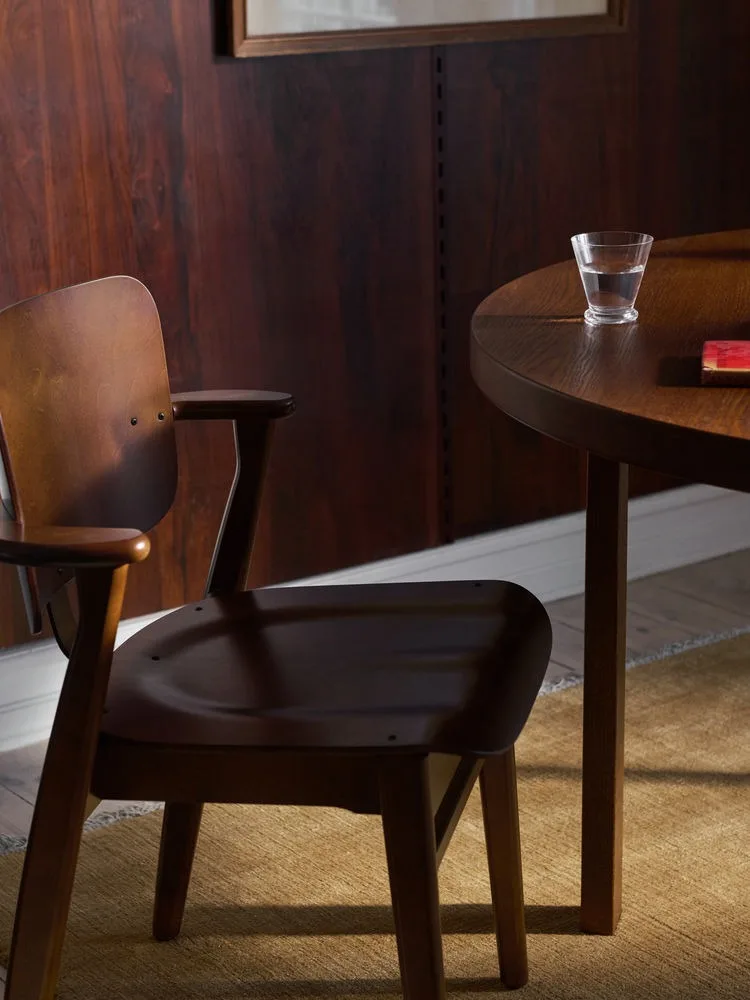 Mørk trestol med armlener ved et rundt bord med et glass vann og en rød bok.