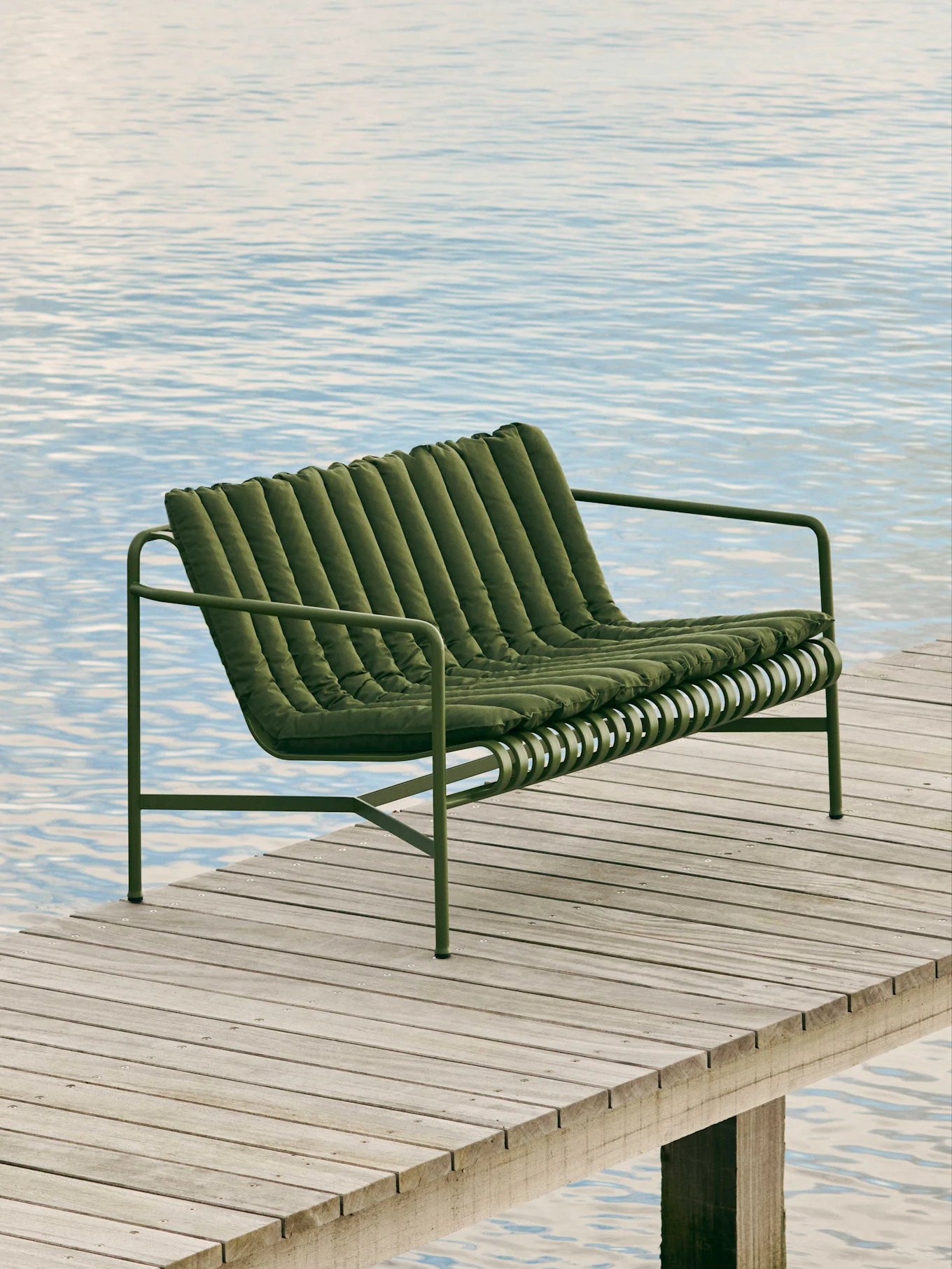 Grønn toseters sofa med polstrede, vertikale puter på en trebrygge over vann.