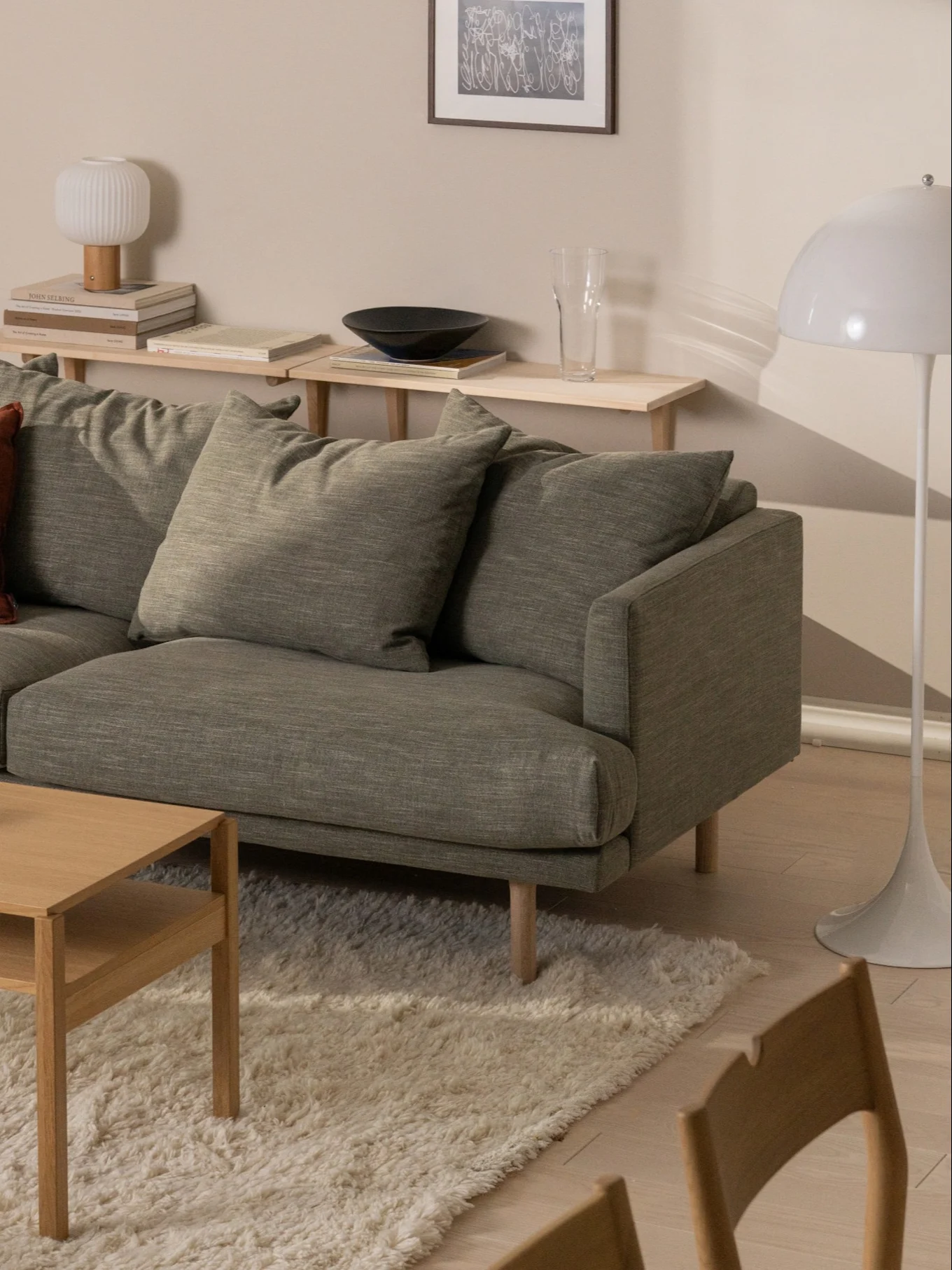 En grønn stoffsofa og et salongbord i lyst tre på et kremfarget teppe i en stue, med hyller og en hvit gulvlampe.