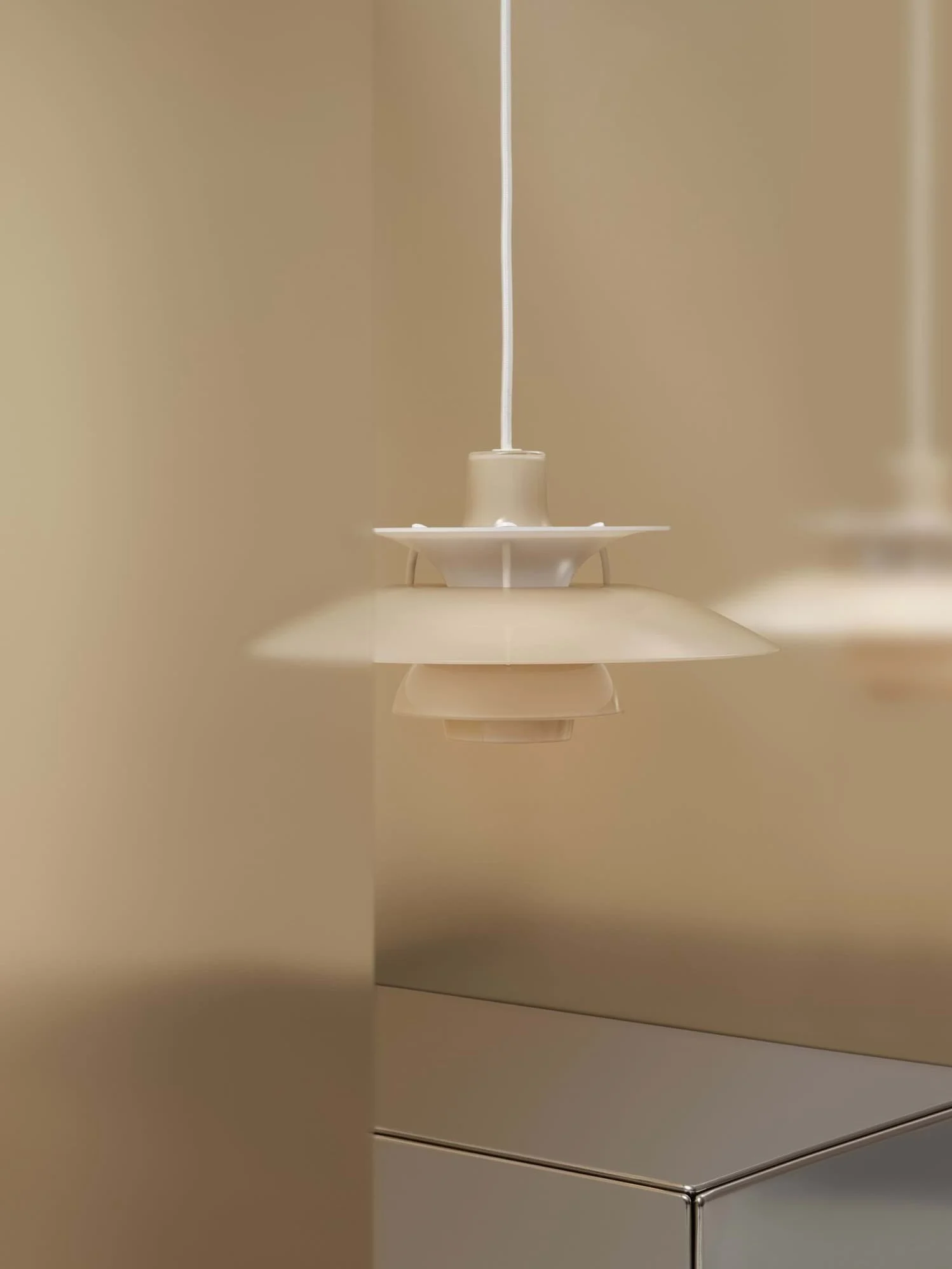 Beige pendellampe med flere skjermer henger foran en beige vegg, med en reflekterende overflate under.