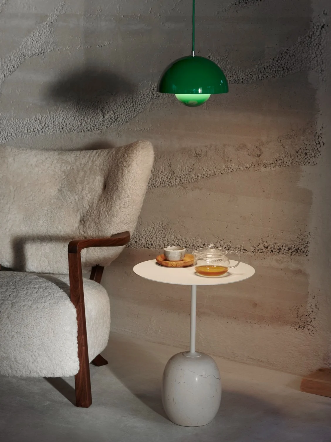 Et koselig interiør med en hvit, lodden lenestol, et sidebord med tekanne og kopp, og en grønn pendellampe over.